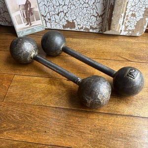 May include: Two antique cast iron dumbbells with a dark, weathered finish. Each dumbbell features a central handle connecting two spherical weights. One of the spherical ends is embossed with the number 2, indicating its weight classification.