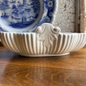 May include: A white ceramic serving dish with a fluted, ribbed exterior and a central raised decorative element featuring a leaf-like motif. The oval-shaped bowl has a smooth, glossy finish and a symmetrical design with curved edges.