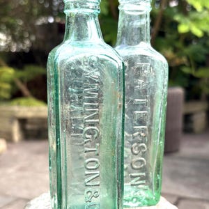 May include: Two vintage light green glass bottles with square bodies and narrow necks. The front bottle features embossed text reading SYMINGTON & CO, while the bottle behind it displays embossed text reading PATERSONS. Both bottles have a clear, translucent finish.
