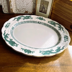 May include: An oval white ceramic serving platter featuring a scalloped edge with a gold rim. The platter is decorated with a green dragon motif and floral patterns along the border, surrounding a central white area with a green geometric band.