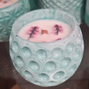 Könnte beinhalten: Eine türkisfarbene Kerze in einem runden, strukturierten Behälter. Die Kerze hat eine weiße Wachsoberfläche mit rosa floralen Akzenten und einen Holzdocht. Der Behälter hat ein gekerbtes Muster.