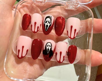 Pode incluir: Um conjunto de unhas postiças com tema de Halloween. As unhas são vermelhas e cor-de-rosa com um design de rosto de fantasma branco e sangue pingando. As unhas são exibidas em uma caixa de plástico transparente.