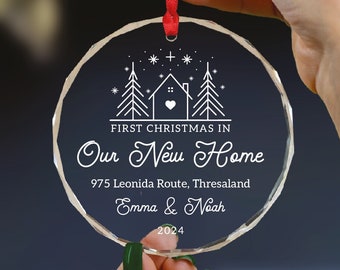 Può includere: Un addobbo natalizio in vetro trasparente con un disegno bianco di una casa e alberi. Il testo "FIRST CHRISTMAS IN OUR NEW HOME" è stampato sull'addobbo, insieme a un indirizzo, nomi e l'anno 2024.