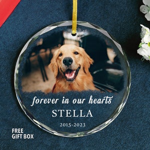 May include: A clear glass ornament with a photo of a golden retriever dog. The ornament has the text "forever in our hearts" and "STELLA" with the dates "2015-2023".