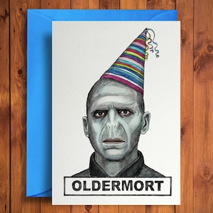 May include: A birthday card with a portrait of a character wearing a party hat with colourful stripes and streamers. The card has the word "OLDERMORT" in a black box at the bottom. The card is inside a blue envelope.
