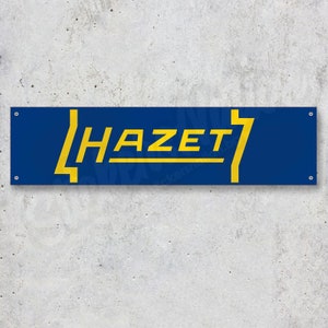Puede incluir: Bandera rectangular azul con la palabra "HAZET" en letras amarillas estilizadas. Un rayo amarillo a la derecha. La bandera está fijada a una pared de hormigón gris.