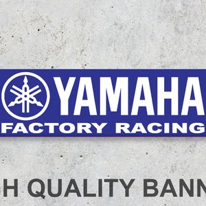 Può includere: Uno striscione blu con testo bianco che recita "YAMAHA FACTORY RACING". Il logo Yamaha è sul lato sinistro dello striscione.