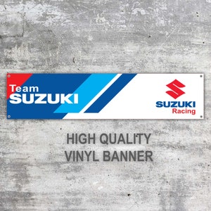 Puede incluir: Una pancarta de vinilo blanca con rayas azules y rojas. La pancarta dice "Team Suzuki" en el lado izquierdo y "Suzuki Racing" en el lado derecho. El logotipo de Suzuki está en el centro de la pancarta.