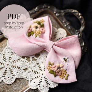 Może przedstawiać: Jasnoróżowa kokarda z haftem kwiatowym i motywem pszczoły. Kokarda jest ozdobiona haftowanymi kwiatami w odcieniach różu, żółci i zieleni. Tekst "PDF step by step instruction" jest widoczny.