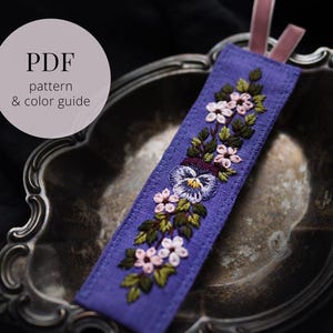 Puede incluir: Un marcapáginas bordado morado con un diseño floral. Presenta flores rosas y blancas, hojas verdes y un pensamiento central. El marcapáginas tiene una cinta rosa y el texto "PDF pattern & color guide" en un círculo blanco.