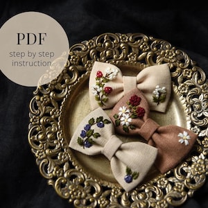 May include: Four embroidered fabric bows with floral designs, including strawberries, daisies, and blueberries, are displayed on a decorative gold-colored plate. The bows are in shades of beige and brown. The image also includes the text "PDF step by step instruction".