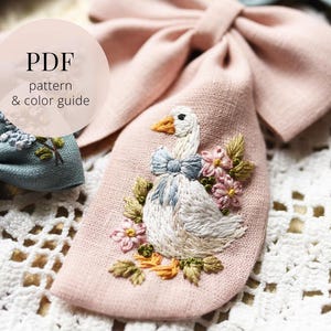 May include: A dusty pink fabric bow featuring hand embroidery of a white goose with an orange beak and a blue neck bow, surrounded by pink flowers and green leaves. The image includes the text PDF pattern & color guide in a circular overlay.
