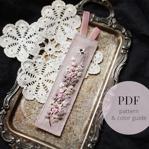 Puede incluir: Un marcapáginas lavanda hecho a mano con flores rosas y blancas bordadas y una cinta rosa. El marcapáginas está sobre una bandeja plateada junto a un tapete de encaje blanco. La imagen incluye el texto "PDF pattern & color guide".