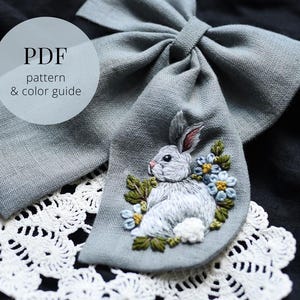 以下が含まれることがあります： 青と黄色の花と緑の葉に囲まれた、詳細な刺繍のウサギが付いた灰色の布のリボン。テキスト「PDF pattern & color guide」は白い円の中にあります。