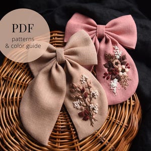 May include: Two fabric bows with embroidered floral designs. The bow on the left is a light brown color and the bow on the right is a light pink color. Both bows have a small bouquet of flowers embroidered on them. The flowers are in shades of pink, white, and brown.