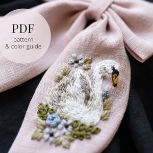 Puede incluir: Lazo de tela rosa claro con un diseño de cisne bordado. El cisne es blanco con pico amarillo, rodeado de bordados florales verdes y azules. El texto "PDF pattern & color guide" está en un círculo blanco.