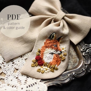 Puede incluir: Lazo de lino beige con un diseño de zorro bordado detallado. El zorro tiene pelaje naranja y blanco, rodeado de flores y bayas. El texto "PDF pattern & color guide" está en un círculo en la esquina superior izquierda.