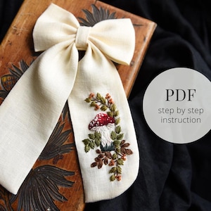 Peut inclure: Un nœud en lin blanc avec un motif de broderie de champignon et de feuillage. Le nœud est sur une surface en bois. Le texte "PDF step by step instruction" est dans un cercle sur le côté droit de l'image.