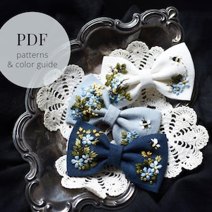 May include: Three embroidered bow ties in white, light blue, and navy blue, each adorned with floral and bee designs. The bows rest on a silver tray with a white lace doily. The image includes text that reads "PDF patterns & color guide."
