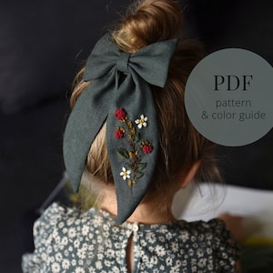 May include: A green fabric hair bow with a floral embroidery design. The embroidery features red berries, white flowers, and green leaves. The bow is tied in a large loop with the ends hanging down. The text "PDF pattern & color guide" is visible in the image.