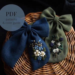 Pode incluir: Dois laços de cabelo de tecido feitos à mão, um azul marinho e outro verde oliva, são exibidos em uma cesta de vime. O laço azul marinho apresenta flores brancas e azuis bordadas. O laço verde tem flores brancas e bagas roxas bordadas.