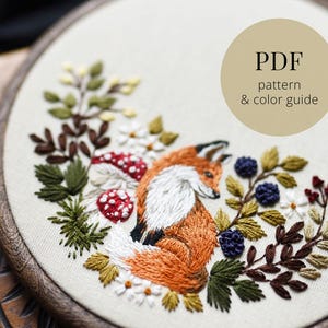 Puede incluir: Diseño de zorro bordado en un aro de madera. El zorro es naranja y blanco, rodeado de hojas verdes, flores blancas, setas rojas y blancas y bayas azules. El texto "PDF pattern & color guide" está en un círculo beige.