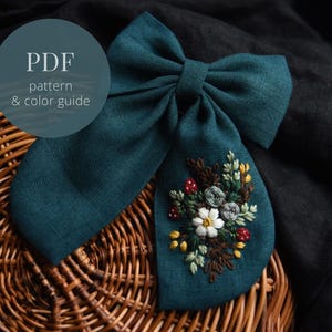 Pode incluir: Um laço de linho azul-esverdeado com um desenho de bordado floral. O bordado apresenta um grupo de flores brancas, bagas vermelhas e folhas verdes. O laço está sobre uma cesta de vime.