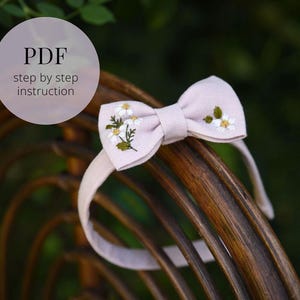 Op de afbeelding: Een roze hoofdband met een strik versierd met witte geborduurde bloemen. De hoofdband ligt op een houten oppervlak. PDF stap voor stap instructie.