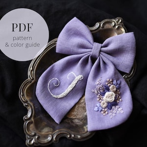 May include: A lilac fabric bow with embroidered floral details and the letter "J". The bow is displayed on a silver tray. The text "PDF pattern & colour guide" is in a white circle in the upper left corner.