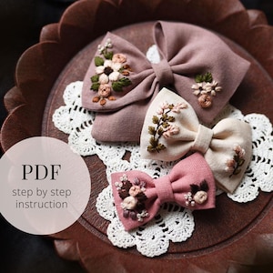 Puede incluir: Tres lazos de tela hechos a mano en tonos rosa y crema, adornados con diseños florales bordados. Los lazos están colocados sobre una bandeja de madera encima de un tapete de encaje blanco. La imagen incluye el texto "PDF step by step instruction".