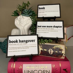 May include: Three rectangular white signs with black frames and text definitions for book, just one more chapter, and book hangover. A white ceramic face planter with green succulents sits behind the signs, placed atop a pink How to Be a Unicorn book.