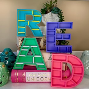 Peut inclure: Des lettres imprimées en 3D colorées épelant "READ" avec des étagères intégrées. Les lettres sont turquoise, violettes, vertes et roses, disposées sur des livres. Des figurines d'oiseaux décoratives et une plante dans un pot blanc en forme de visage sont également présentes.