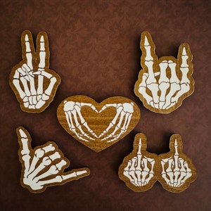 May include: Five wooden skeleton hand cutouts, each with a different design. The designs include a peace sign, a heart, a rock and roll hand gesture, and two thumbs up. The cutouts are painted white on a brown background.