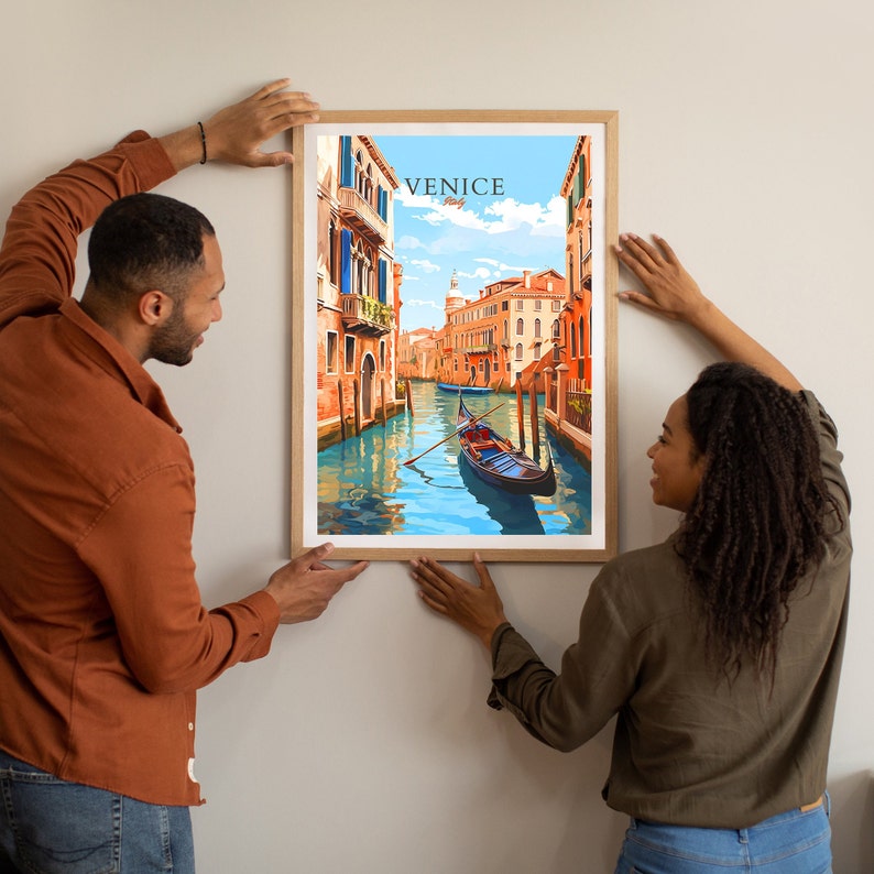 Puede incluir: Una pareja cuelga una impresi&oacute;n enmarcada de una ilustraci&oacute;n colorida de una g&oacute;ndola en un canal de Venecia, Italia. La impresi&oacute;n tiene el texto "VENICE Italy" en la parte superior.