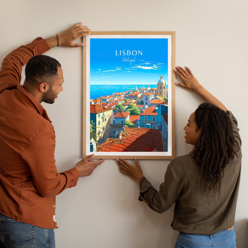 May include: Two people are hanging a framed print of a colorful illustration of Lisbon, Portugal. The print shows a cityscape with red-tiled roofs and a blue sky.