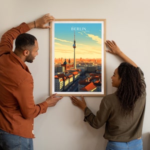 May include: A couple hangs a framed print of a cityscape with a tall tower in the center. The print is titled "BERLIN" and features a sunset sky with a light blue and orange color scheme.