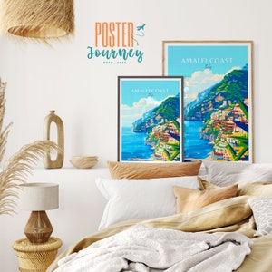 May include: Two framed posters with a colorful illustration of the Amalfi Coast in Italy. The posters are hanging on a white wall above a bed with white bedding and brown pillows.