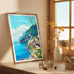 May include: A framed poster with a colorful illustration of the Amalfi Coast in Italy. The poster features a vibrant blue sea, a rocky coastline, and colorful buildings built into the hillside. The text "Amalfi Coast Italy" is printed in white at the top of the poster.