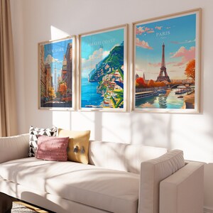 May include: Three framed travel posters with colorful illustrations of New York City, the Amalfi Coast, and Paris. The posters are hanging on a white wall above a white couch with pillows.