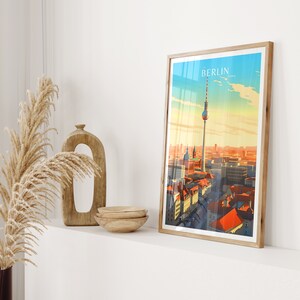May include: A framed print of a cityscape with the Berlin Television Tower in the center. The print has a vintage travel poster aesthetic with a blue sky and orange and red buildings.