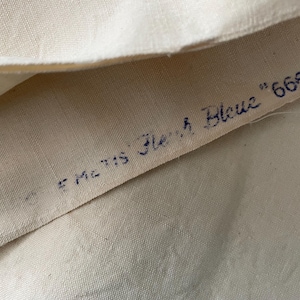 May include: Close-up of a white fabric with the text "E Mc 115 Flesh Blue" and "665" written in blue ink.