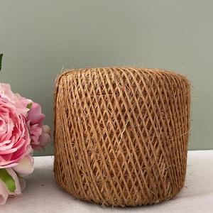 Può includere: Un grande rotolo di spago marrone naturale, realizzato con fibre naturali, con un motivo intrecciato. Lo spago è avvolto strettamente attorno al rotolo.