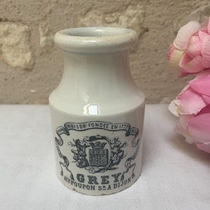 Puede incluir: Un pequeño tarro de cerámica blanquecina con la parte superior redondeada y un cuerpo cilíndrico. El tarro presenta un emblema gris con el texto "MAISON FONDEE EN 1777" y "GREY A. POUPON S.A. DIJON". El fondo es una pared clara y texturizada.