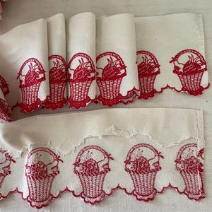 Könnte beinhalten: Weißer Stoff mit roten gestickten Obstkörben. Der Stoff hat einen gewellten Rand.