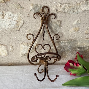 Peut inclure: Applique murale de style antique en finition bronze. La ferronnerie présente des détails en volutes, une petite étagère et deux crochets. L'applique est fixée sur un mur en pierre texturée. Une fleur rouge et verte est au premier plan.