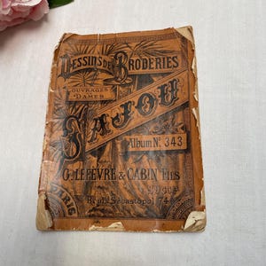 May include: An antique book cover with the title "Dessins de Broderies" in black script. The cover is a faded orange color with decorative floral and geometric patterns. The book is titled "LAFOU" and "Album No. 343".