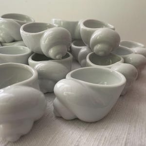 Può includere: Una collezione di ciotole bianche in ceramica a forma di conchiglia. Le ciotole sono impilate, mostrando la loro finitura lucida e gli intricati disegni a spirale. Le aperture delle ciotole sono visibili, suggerendo che siano progettate per contenere piccoli oggetti.