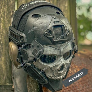 May include: A black tactical helmet featuring a silver skull-shaped face mask with dark eye lenses. The helmet includes side-mounted tan ear protection, a top-mounted rail system, and a front-facing mount. A black tag with the white text NOMAD is attached.