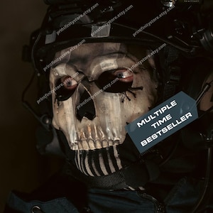 Puede incluir: Primer plano de una máscara de calavera con ojos rojos, parte de un casco táctico. La máscara tiene un diseño esquelético con la parte inferior de la cara a rayas blancas y negras. Incluye el texto "MULTIPLE TIME BESTSELLER".