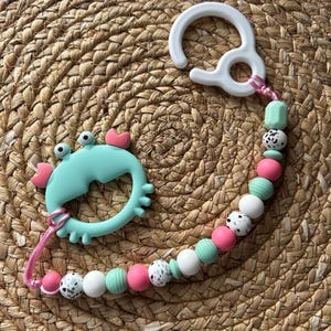 May include: A baby's teething toy and pacifier clip. The teether is crab-shaped in light blue with pink details. The clip has a string of pink, white, mint green, and speckled beads. The clip is white.